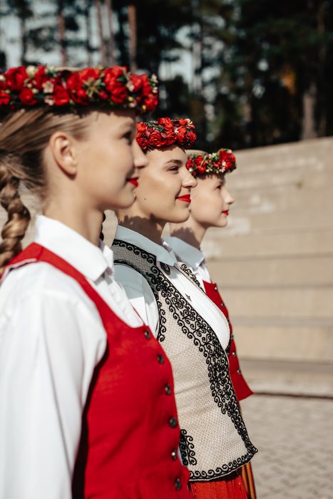 Trīs māsas tautastērpos ar sarkaniem ziedu vainagiem profilā pie Mežaparka estrādes