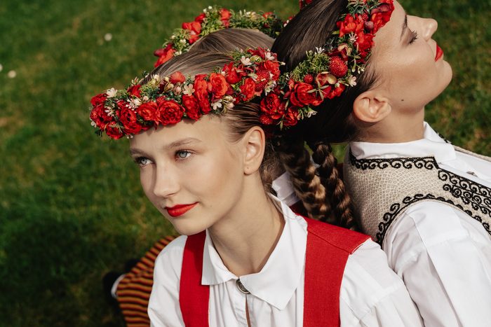 Divas māsas tautastērpos ar sarkaniem vainagiem mugura pret muguru Mežaparkā