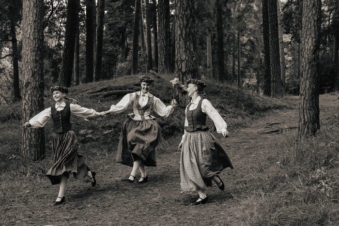 Melnbalta fotogrāfija: trīs māsas skrien pa priežu mežu Mežaparkā, sadevušās rokās