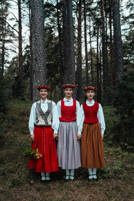 Trīs māsas pilnā augumā tautastērpos priežu mežā Mežaparkā