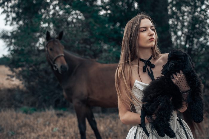 Sieviete gaišā kleitā ar melnu lentīti blakus zirgam
