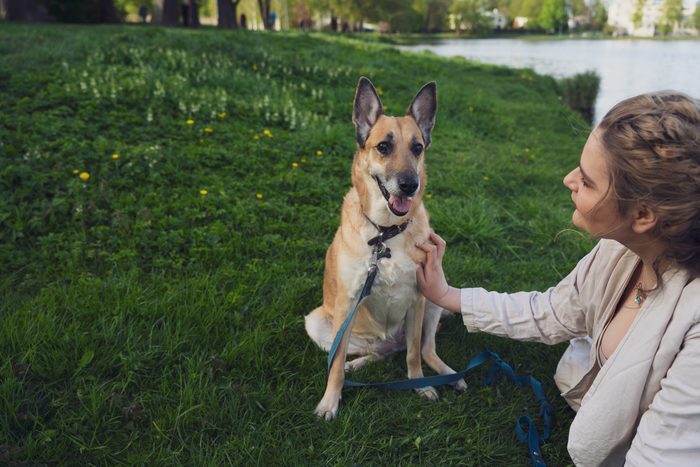 Sieviete gaišā kleitā ar suni un ceriņiem parkā