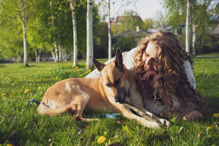 Sieviete gaišā kleitā ar suni un ceriņiem parkā