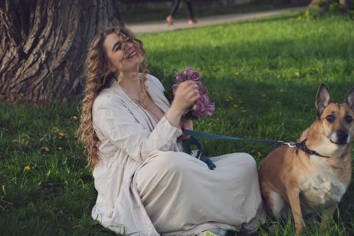 Sieviete gaišā kleitā ar suni un ceriņiem parkā