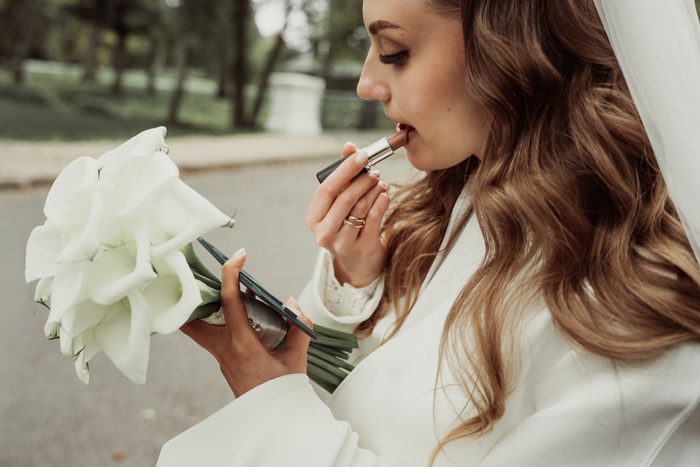 The bride applying lipstick in Ķemeri Park, holding a white calla bouquet