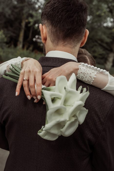 The bride hugging the groom from behind, calla bouquet against his jacket, in Ķemeri