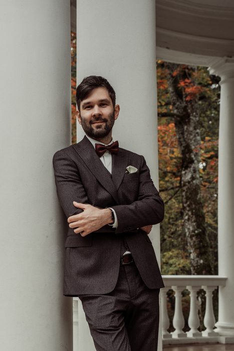The groom with his arms crossed by a column, autumn leaves behind him, in Ķemeri