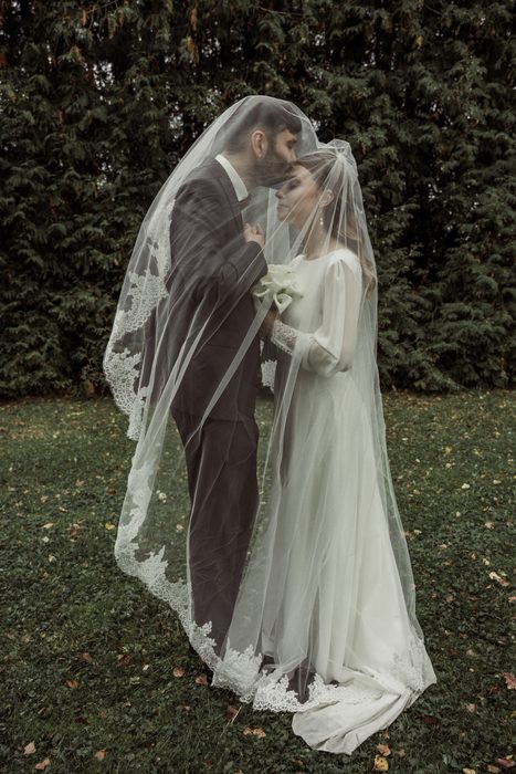 The couple under the long lace veil, the bride looking at the camera, in Ķemeri