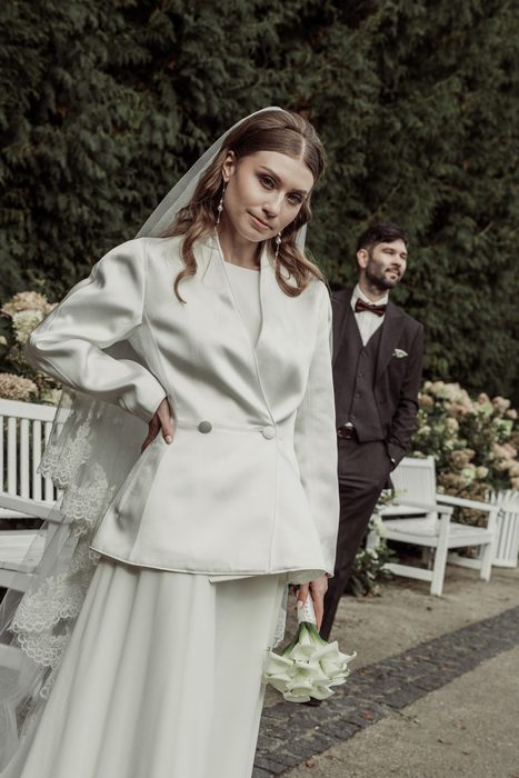 The bride in a white jacket, the groom behind her by the white benches in Ķemeri